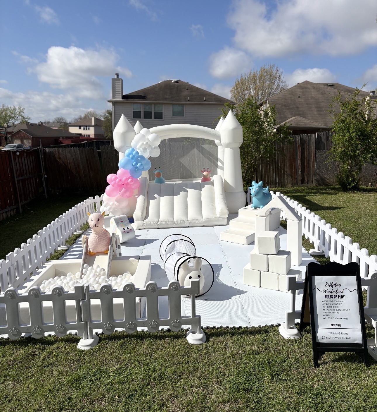White bounce house with pink and blue balloons and fenced soft play area