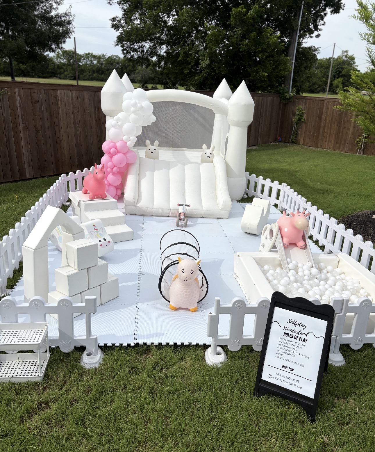 White bounce house with white balloon garland, ball pit, slide and soft play toys outdoors