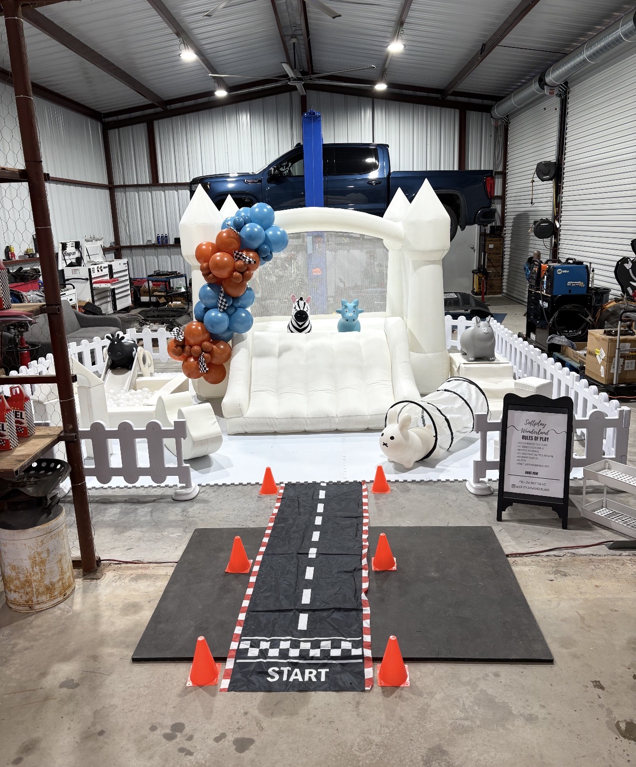 White bounce house with blue and orange balloon garland and race-track soft play